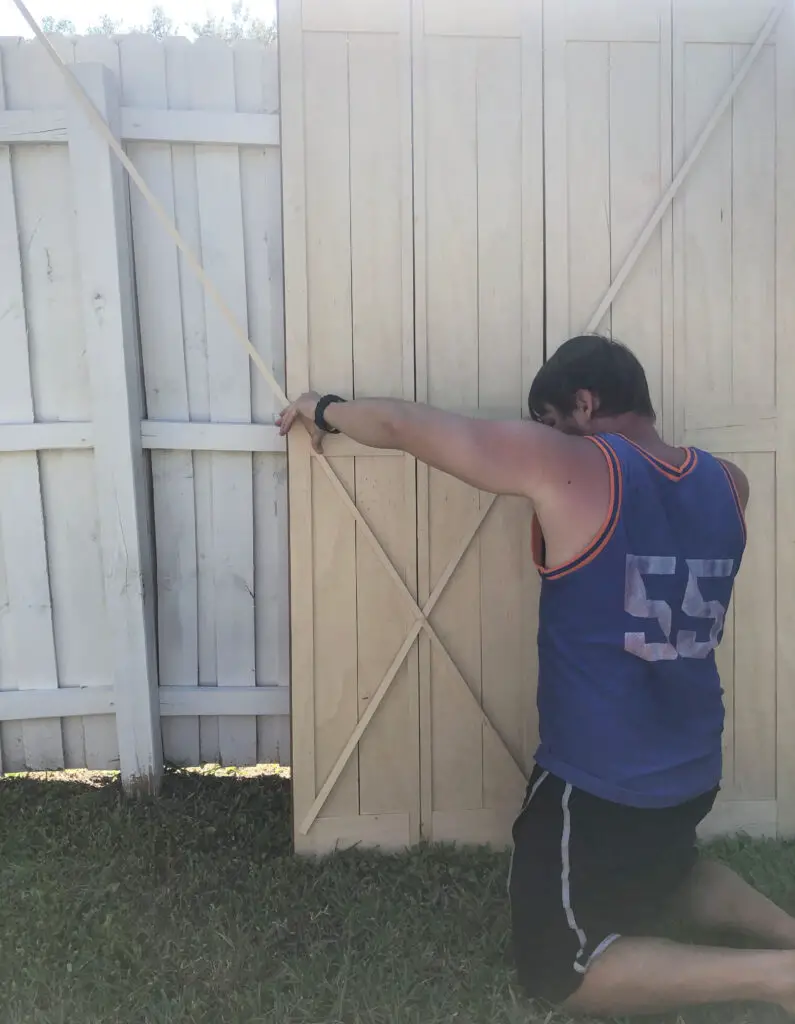 man holding wood pieces into an x to decide on barn door design. The barn door is leaned up against a white fence