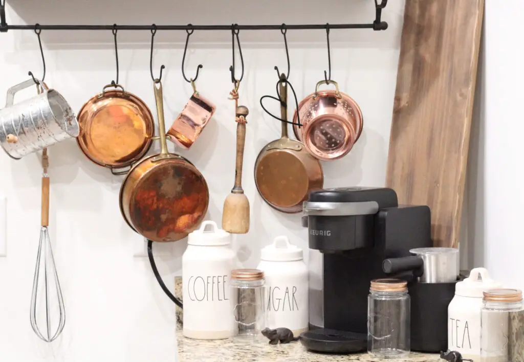 copper pots hanging on a black metal shelf rod with a keriq coffee maker and white canisters. 