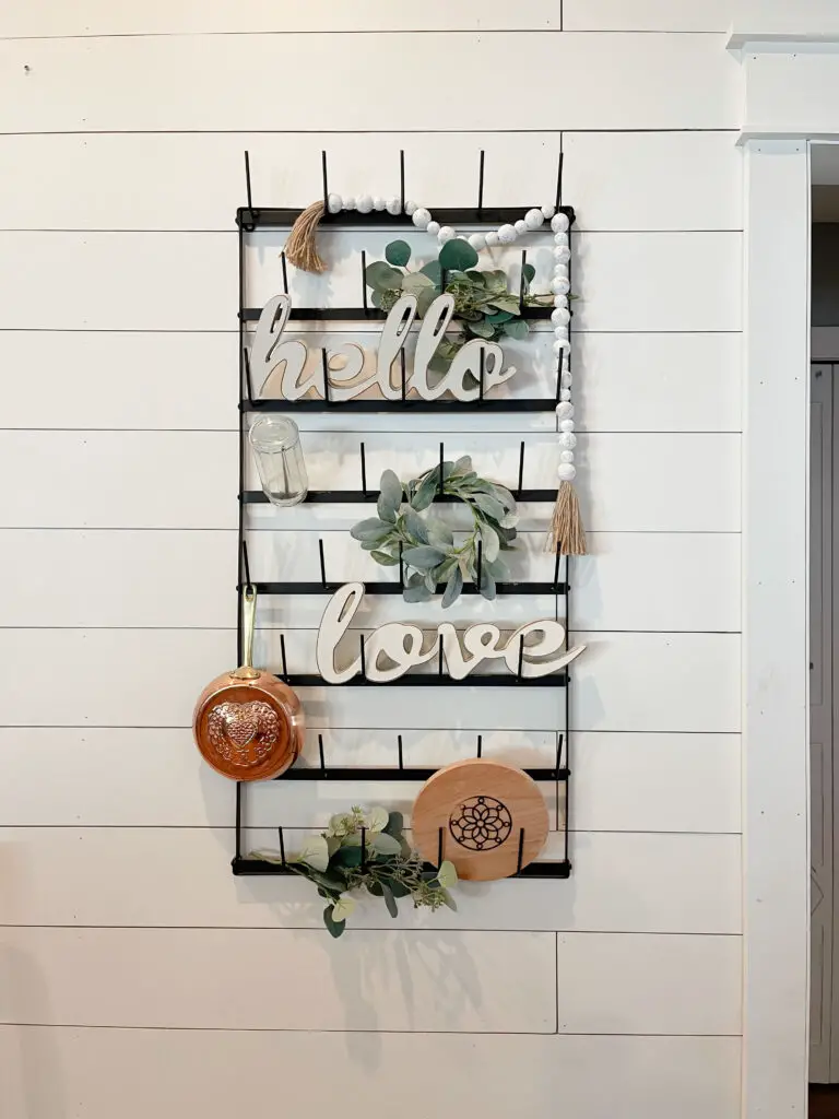black metal rack hanging on a white shiplap wall with greenery, and a copper pan, wood circle, and hello love written in white wood hanging in a farmhouse dining room