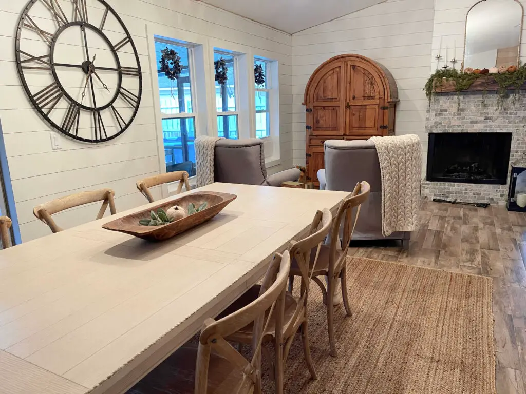 farmhouse dining room with an ivory dining farmhouse table and wood grain crossback chairs, a large metal clock on the wall and white shiplap walls 