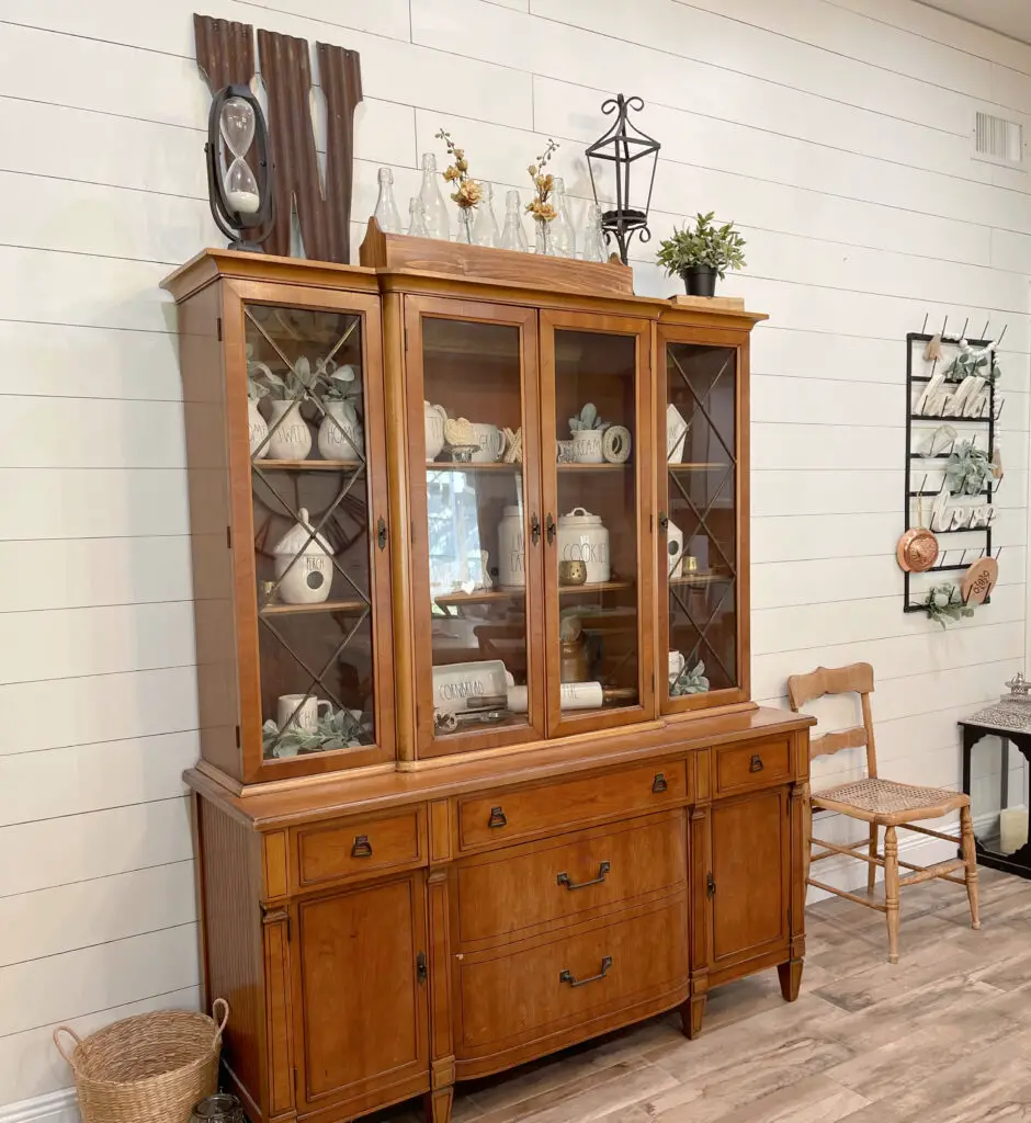 wood toned hutch with rae dunn pieces inside and greenery against a white shiplap wall in a farmhouse dining room