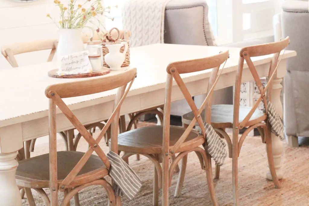 an ivory farmhouse table with wood cross-back chairs and cloth napkins hanging from each chair that are gray and white. 