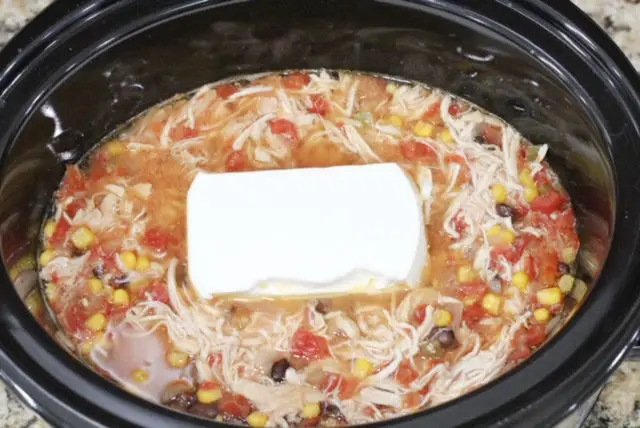 a close up of a black crockpot with shredded chicken, black beans, corn, onion, chicken broth, and a block of cream cheese in it