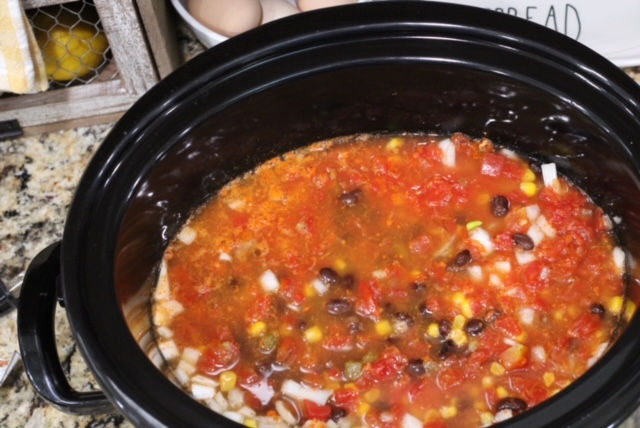 a close up of a black crockpot with corn, black beans, onion, chicken broth in it