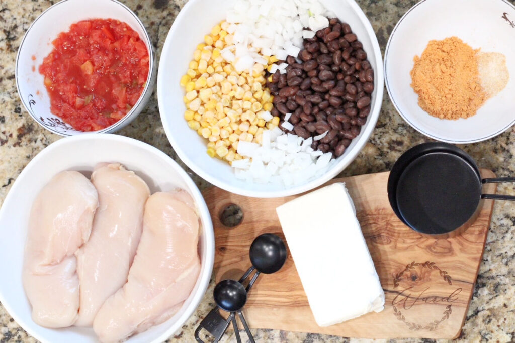 close up of white bowls on a granite countertop with various ingredients such as chicken breasts, corn, onion, black beans, taco seasoning