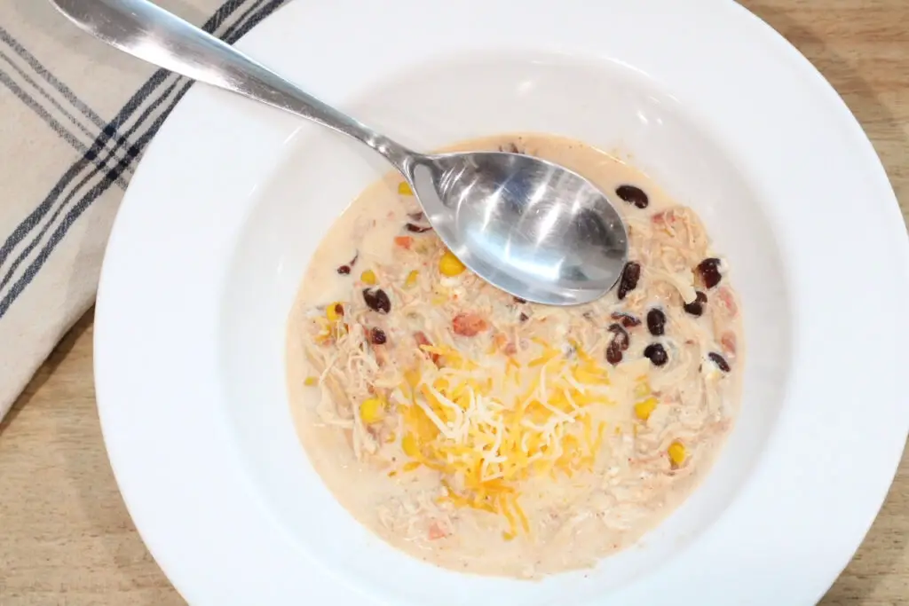 close up of a white bowl with creamy taco soup and a silver spoon and white and blue towel on a cutting board. 