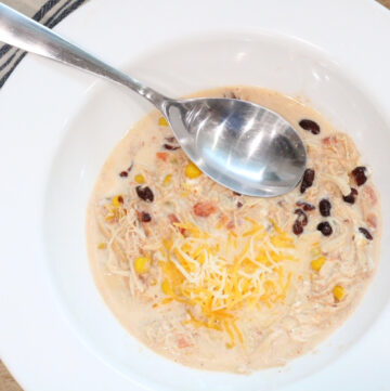 close up of a white bowl with silver spoon, cheese, black beans, corn, onion inside, on top of a wood cutting board