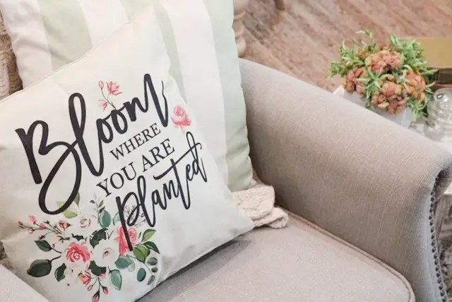 A pillow cover that says Bloom where you are planted with spring florals on it and a white and green striped pillow behind it on a grey accent chair