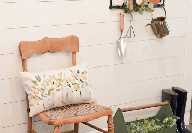 a wooden antique chair with a yellow lemon pillow against a white shiplap wall and various spring décor hanging on  black hooks, a green caddy with greenery on the ground next to navy galoshes 