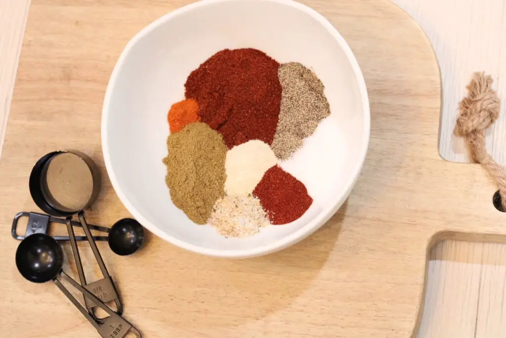 a close up of a white bowl with mounds of 8 different seasonings in it: paprika, salt, pepper, garlic salt, cayenne pepper, chili powder, cumin and black measuring spoons beside the bowl on top of a wood cutting board