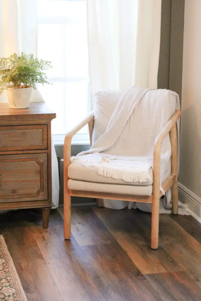 beige accent chair with wood trim on dark wood floors next to a green plant on a brown wood nightstand and both are in front of a window