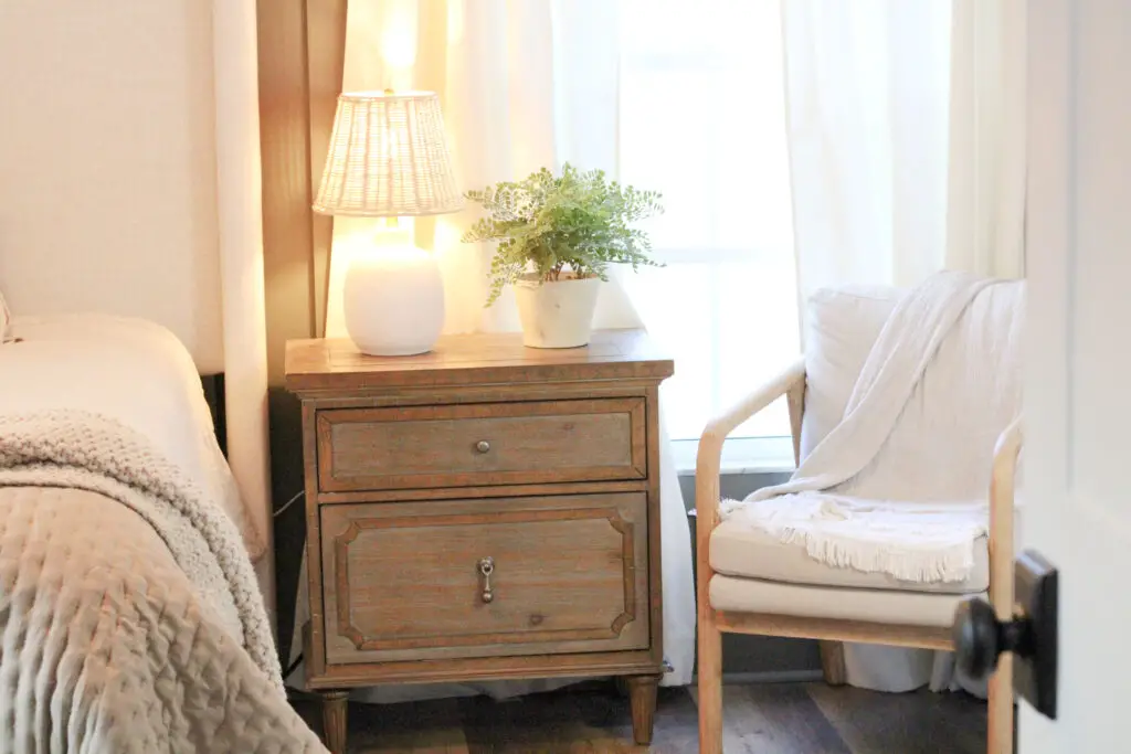 brown wood nightstand with white table lamp and green plant on top with a beige accent chair beside it in a master bedroom