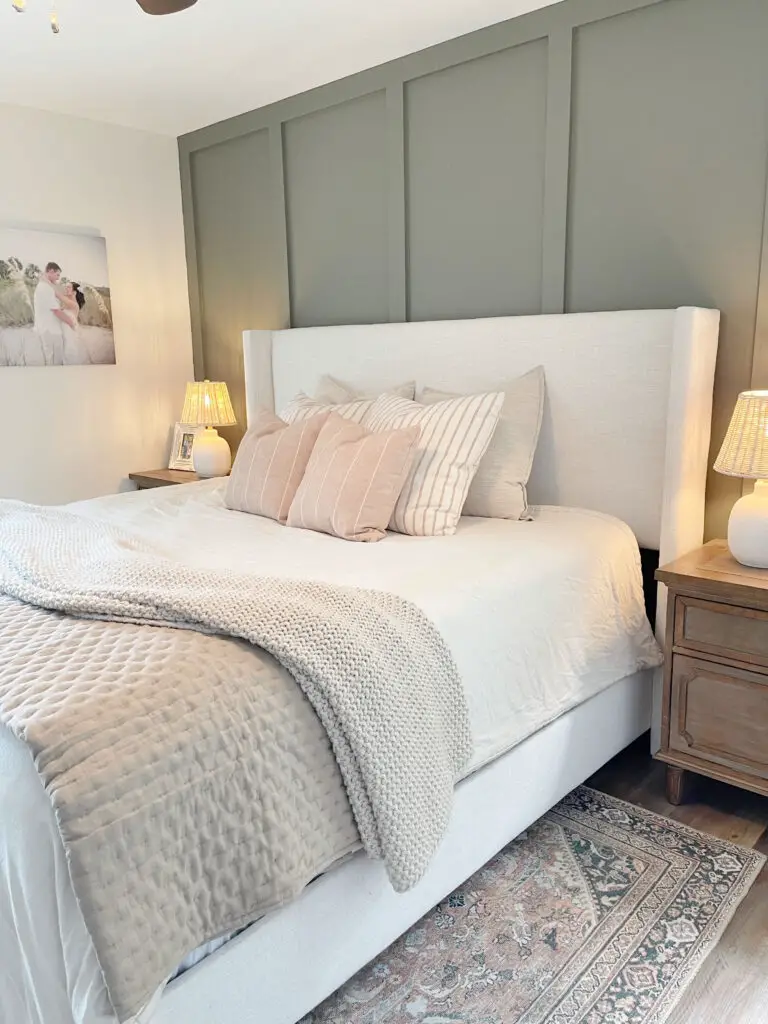 Green board and batten accent wall with linen white bed in front of it. With white and orange throw pillows on the bed and beige throw blankets. 