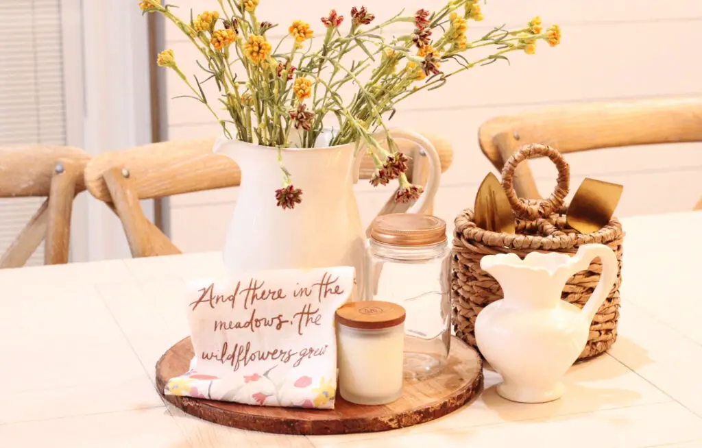 Close up of a white pitcher with yellow and red spring florals in it on a wood service board, next to a wicker basket and a small white pitcher in front, all on a white dining table. 