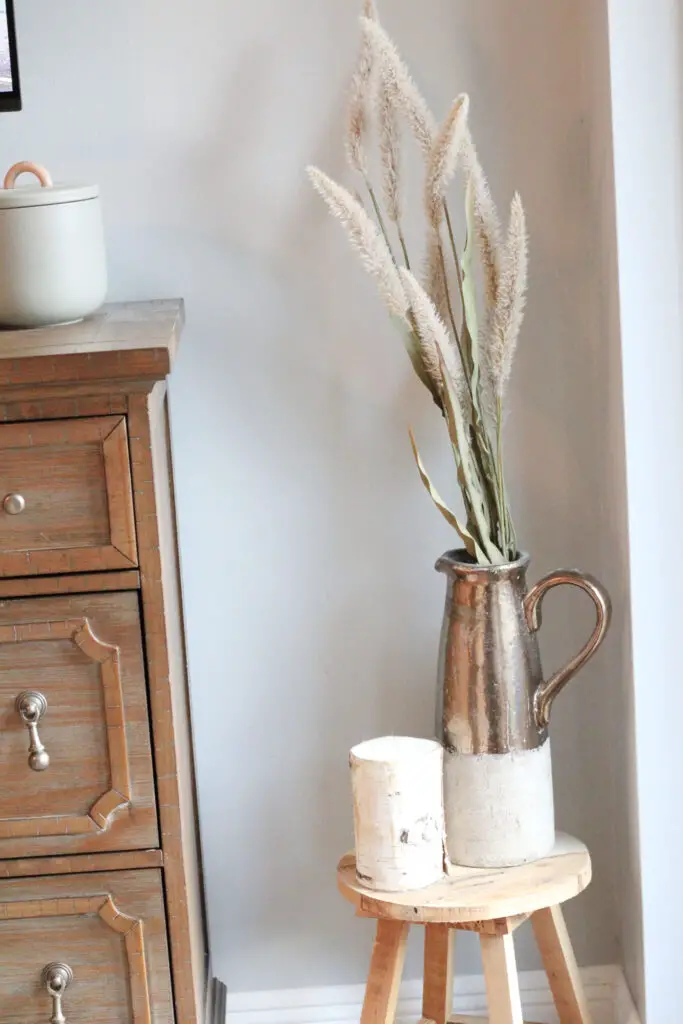 Close up of a gold and beige vase with willow stems in it on a wood stool with a birch candle in front of it. 