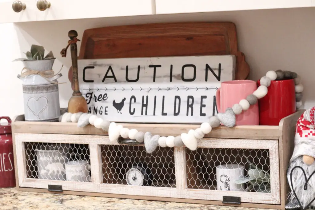 close up of a shelf with a pink and red mug, a white sign, and a garland of white and gray hearts. 
