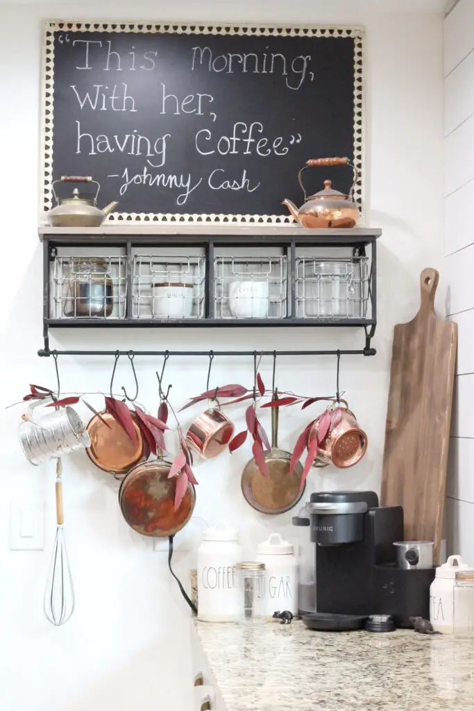 A shelf on the wall with a holder with coffee in it, a holder with sugar in it, and copper antique pots hanging off of the shelf. 