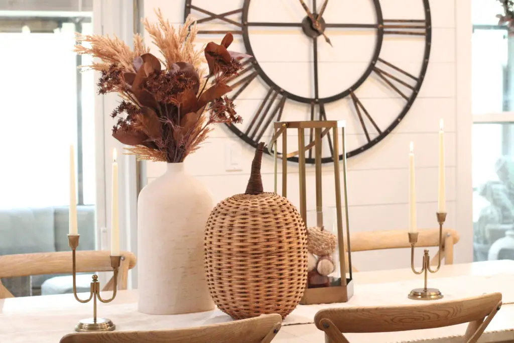 A close up of a white vase with brown and beige fall florals and a large wicker pumpkin in front of the vase, with gold brass candleholders with white candles lit on either side of the vase and pumpkin, all on a white dining table. 