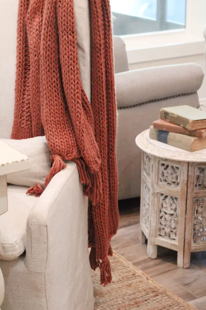 close up of a rust colored knit throw blanket thrown over a white linen colored dining chair, with beige side table with antique books on top. 