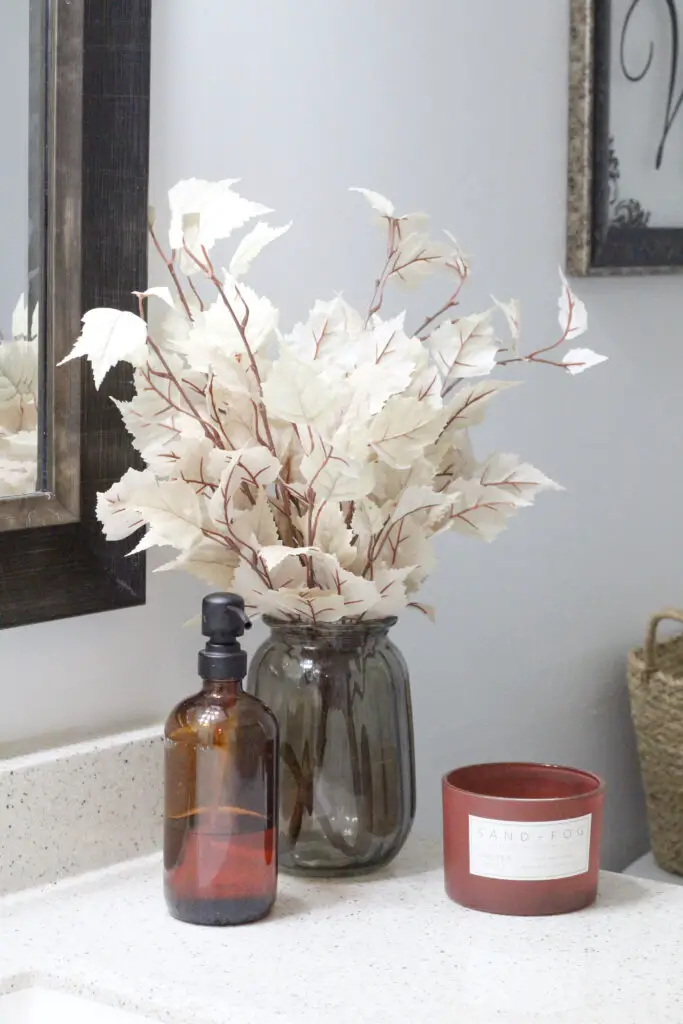 Close up of a dark green vase with white fall leaves in it next to an amber colored soap dispenser on a bathroom counter