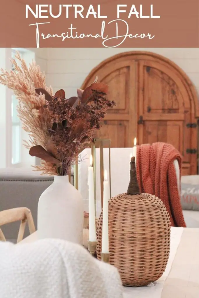 a close up of a white vase with brown fall stems and white candlesticks that are lit and a wicker pumpkin on a white dining table. The words "Neutral Fall Transitional Decor" at the top of the image 