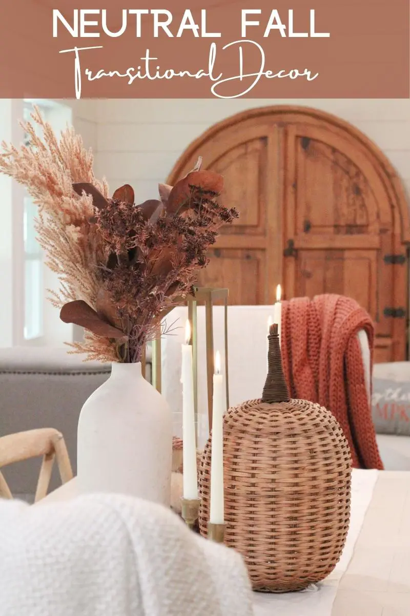 a close up of a white vase with brown fall floral stems and a wicker pumpkin in front of it, with white tapered candles that are lit on a white dining table, and a wood colored arched armoire in the background. The words "Natural Fall Transitional Decor" written at the top