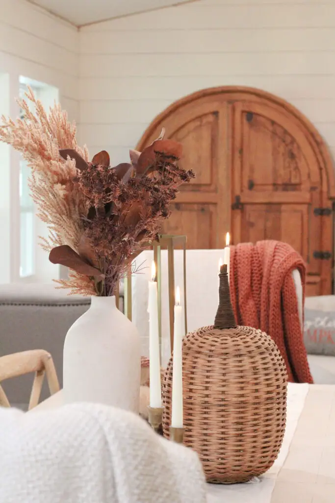 A close up of a white vase with brown and beige fall florals and a large wicker pumpkin in front of the vase all on a white dining table. 