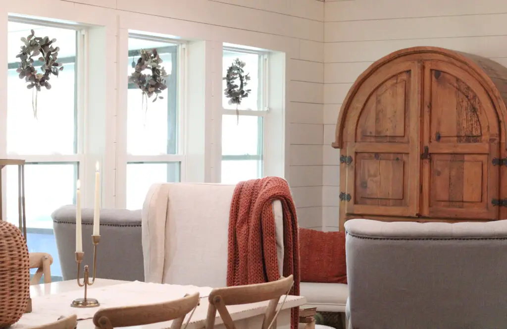 A dining room with white shiplap walls and 3 farmhouse framed windows with green wreaths in them and a white dining table with a white linen dining chair and rust colored knit throw over the chair. 