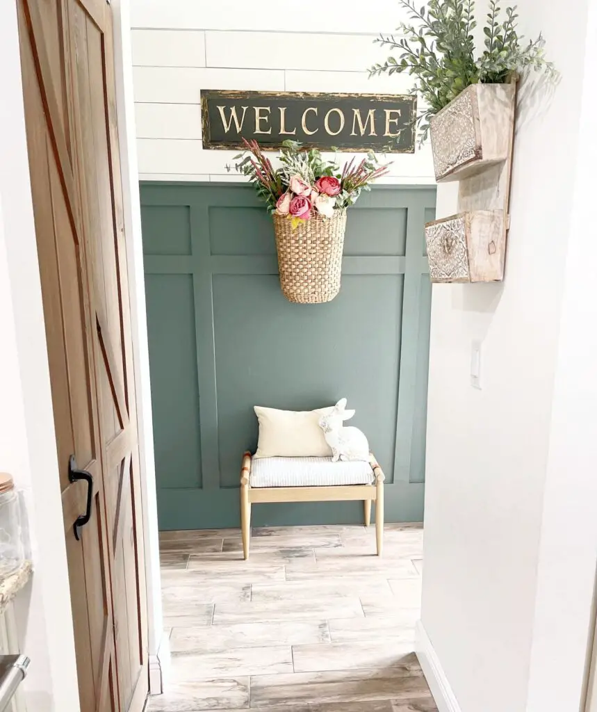 Close up of a green board and batten wall with white shiplap above it and a basket with pink flowers hanging on the wall and a bench underneath. 