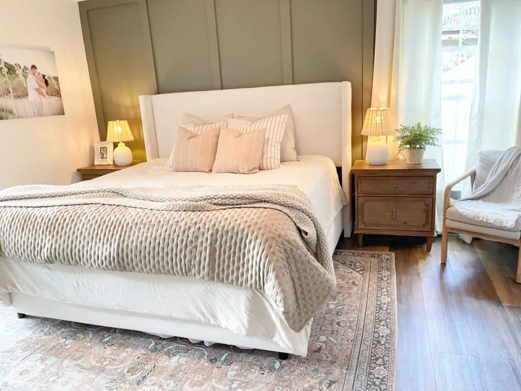 A white linen bed against a dark green board and batten wall with a wood colored night stand and lamp on it and plant.