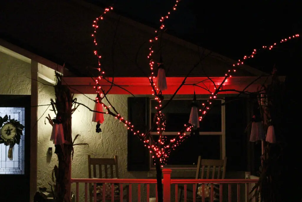 Close up of a black tree at night with orange lights lit up and white ghosts hanging from the tree. 