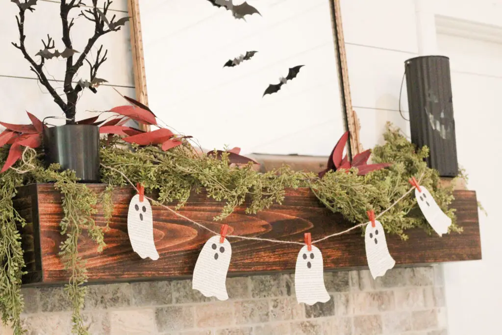 Close up of a wood fireplace mantel and a garland of white book paper ghosts hanging on a burlap string with orange clothes pins. 