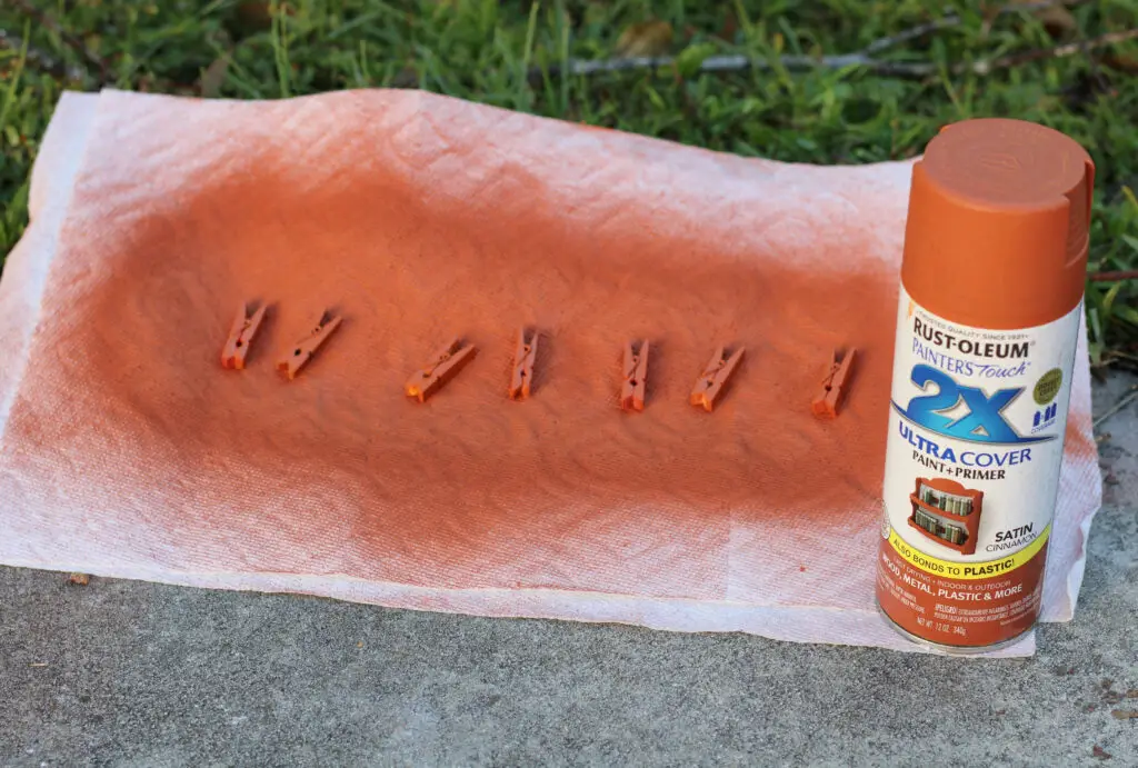 Close up of tiny clothes pins on a white paper towel covered in orange spray paint with the can of orange spray paint next to it on the green grass. 