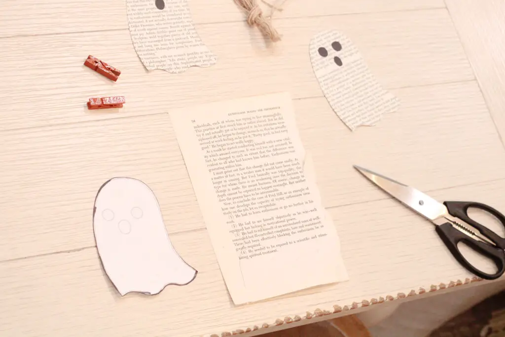 Close up of a white paper ghost outline, a book page, a black scissors, 2 orange clothes pins, burlap string, and a book paper ghost on a white table.