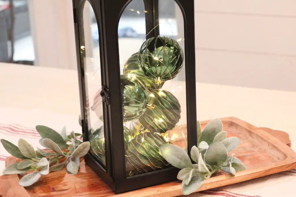 Close up of a black lantern with green and gold Christmas ornaments and fairy lights that are on inside on top of a wood colored tray with some greenery stems on it. 