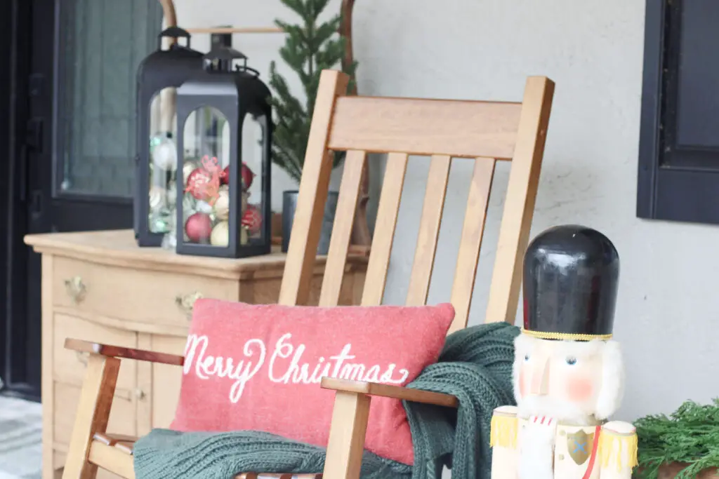 a wood colored dresser with two black lanterns on top of it with green and red and gold Christmas ornaments inside, a wood colored rocking chair in front with a red pillow that says Merry Christmas on it and a green throw blanket underneath, beside it is a large wooden nutcracker all on a front porch. 