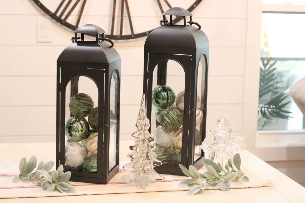Close up of two black lanterns with gold and green Christmas ornaments inside them, on a white dining table with a glass Christmas tree and Santa in front. 