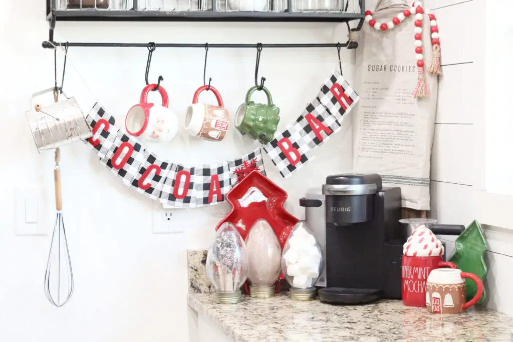 Close up of a a sign that says cocoa bar and has Christmas mugs hanging on a black wire shelf. With glass containers with different ingredients in it. Hot chocolate mix, marshmallows, and chocolate covered spoons inside. 