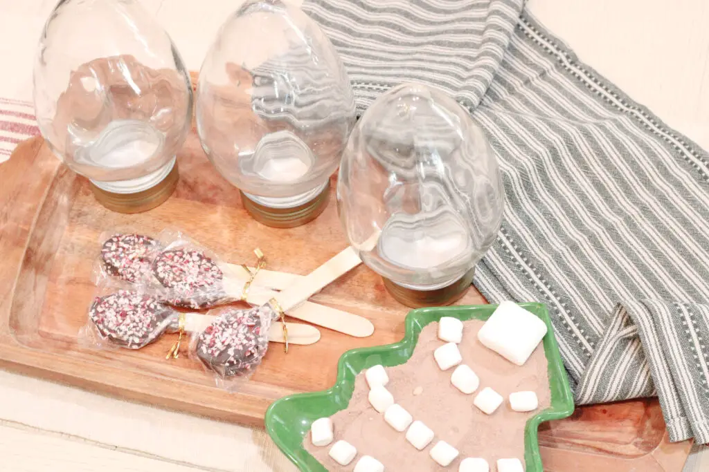 A close up of three glass containers in the shape of large Christmas lights, some chocolate covered wood spoons, and a green Christmas tree plate with hot chocolate mix and marshmallows. 