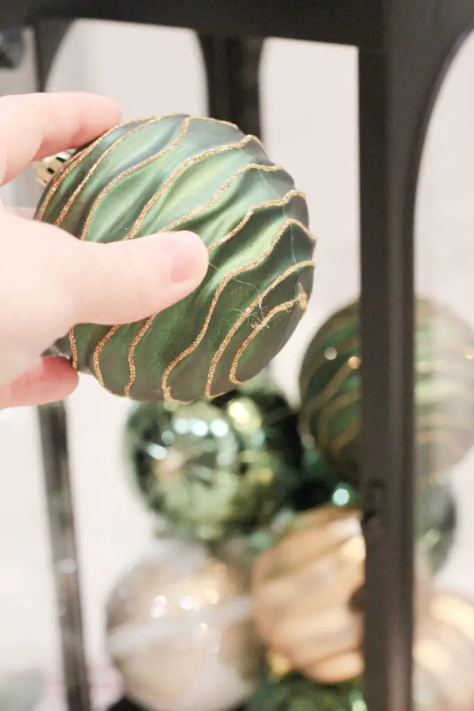 Close up of a hand holding a green Christmas ornament and putting it into a black lantern with other green and gold Christmas ornaments inside.
