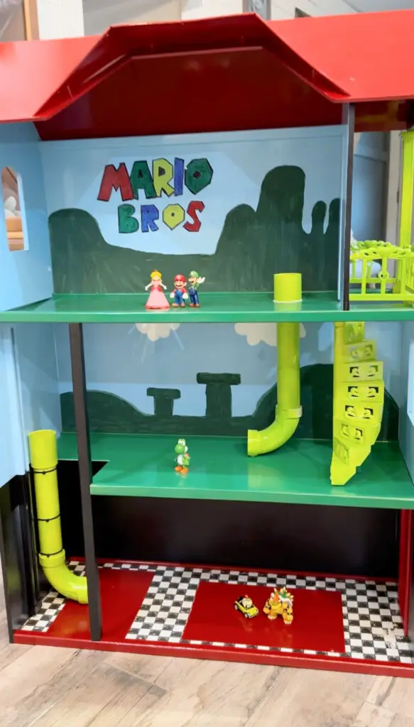 Close up of the Mario playhouse that has a red roof, blue back wall, green floors, and Mario plastic characters inside. 