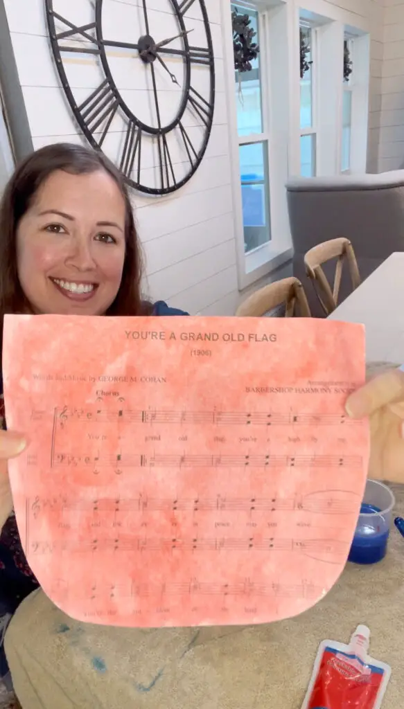 A woman holding up a close up of a red painted piece of paper with the song on it "You're A Grand Old Flag" 