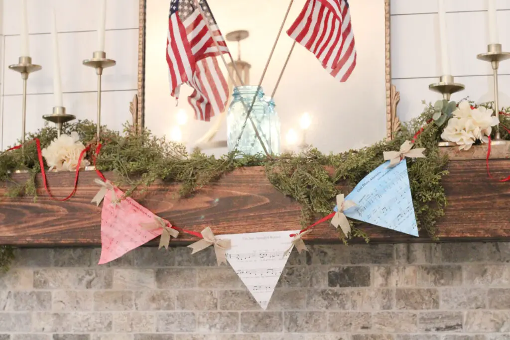 Close up of a red, white, and blue colored garland that has red string and the sheets of paper have black music notes on them. It is hung on a wood colored mantel with greenery. 