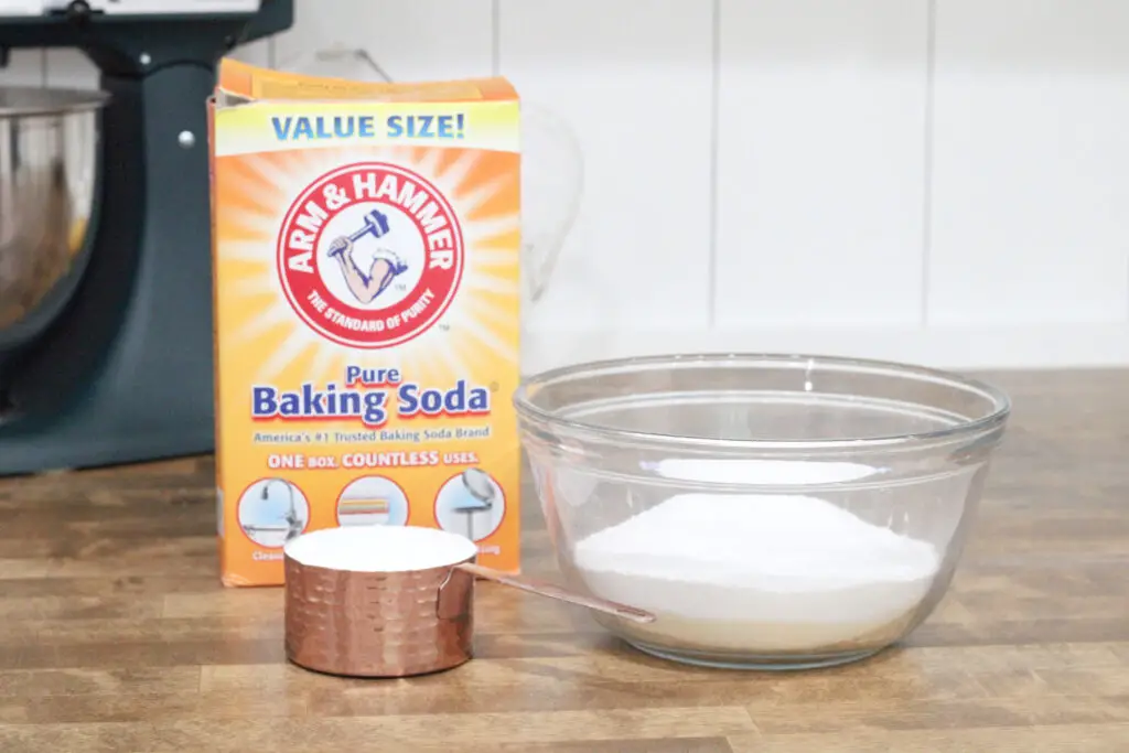 close up of a copper cup measuring cup with white powder in it, a glass bowl with white powder in it, a yellow box of baking soda