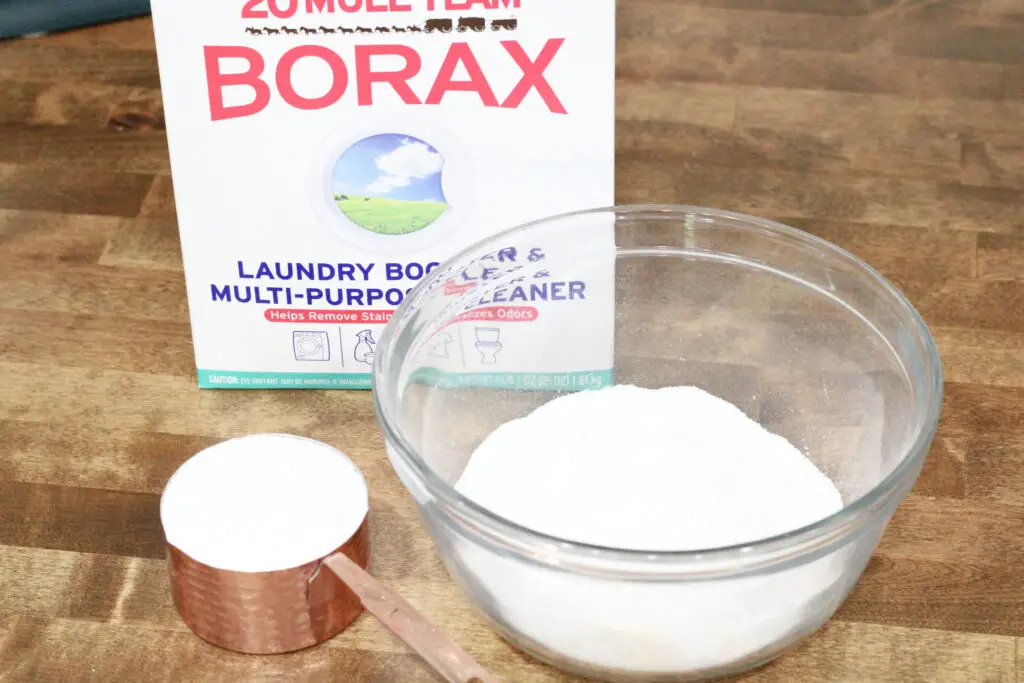 Close up of a copper cup measuring cup with white powder in it, a glass bowl with white powder in it, and a white box of borax
