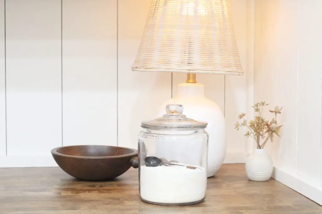 a glass container with lid with white powder inside, a wood antique bowl, a white lamp with wicker shade, on a wood countertop