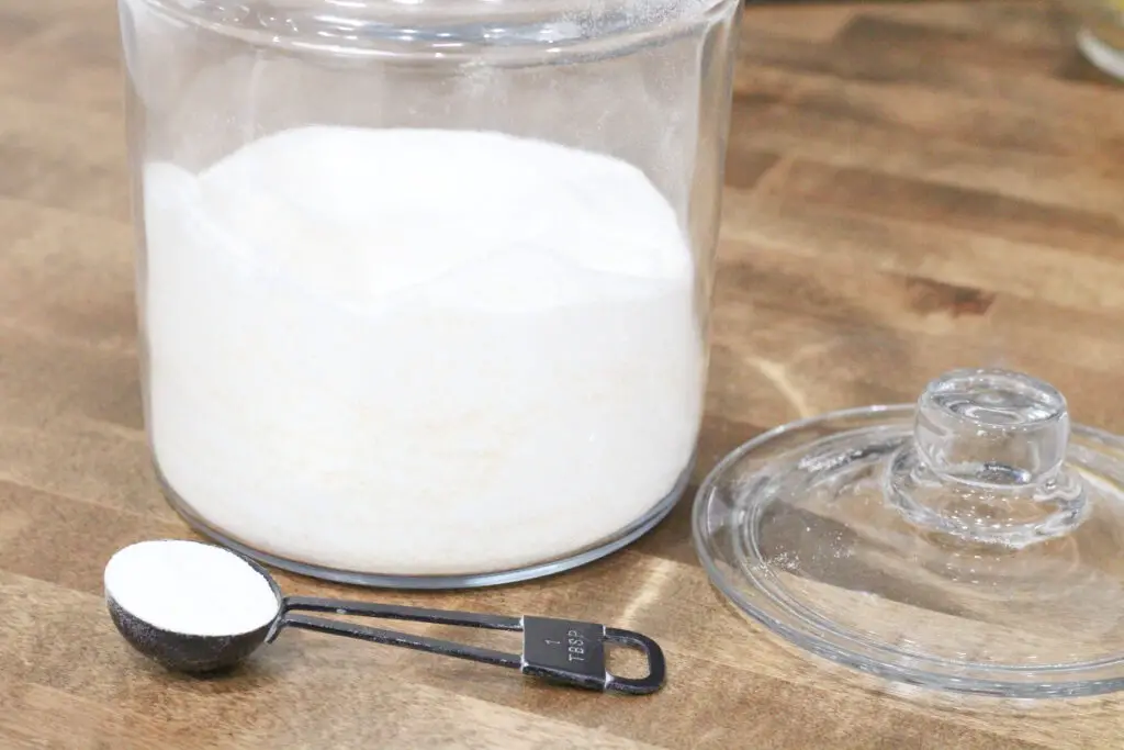 close up of a glass container with lid with white powder in it and a black tablespoon measuring spoon with white powder in it. 