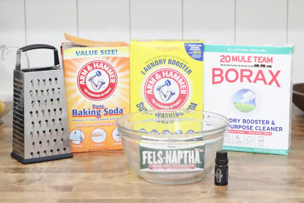 close up of a cheese grator, a box of baking soda, a box of washing soda, a box of borax, a glass bowl, a bar of soap, and essential oil jar. 