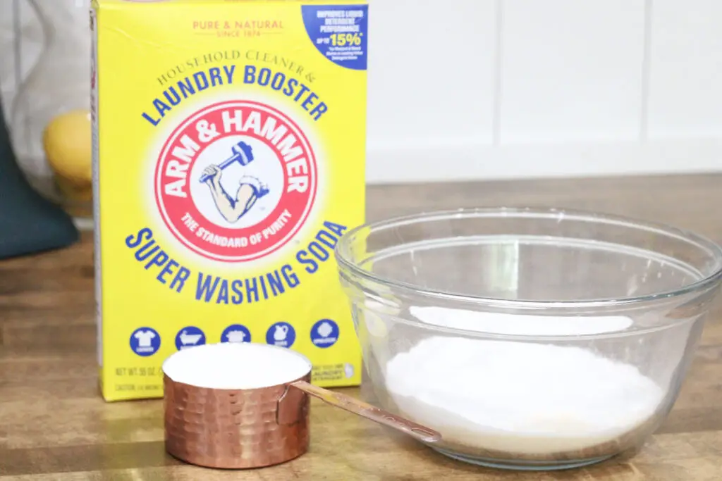 Close up of a copper cup of white powder, a glass bowl with white powder, and a yellow box of super washing soda