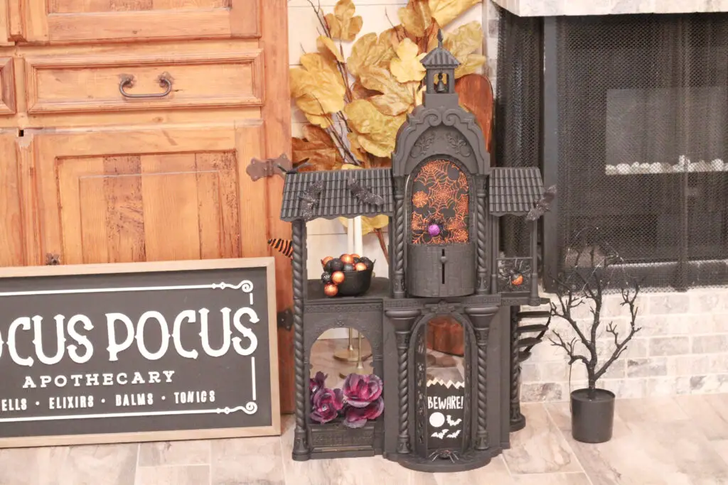black Halloween house with purple flowers, orange spider webs, black and purple spider, a black tree next to the house, all in front of a wood cabinet